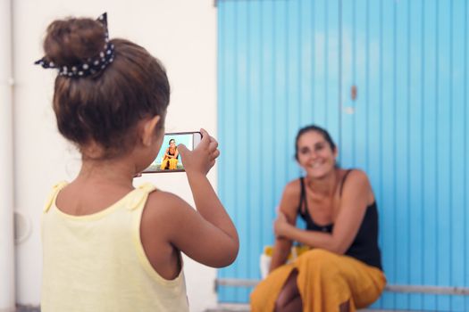 little girl takes photos with the smart phone to her mother in front of a blue door, single-parent family on vacation, divorced woman enjoying with her daughter