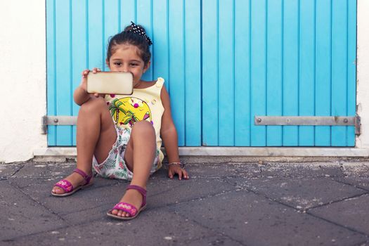 little girl takes a selfie with the smart-phone in front of a blue door, girl enjoying with the technology on vacation
