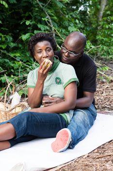 love couple sitting on a mat in the bush. the man sitting behind his wife, looks her eat an apple.