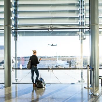 Young casual female traveler at airport, holding smart phone device, looking through the airport gate windows at planes on airport runway.