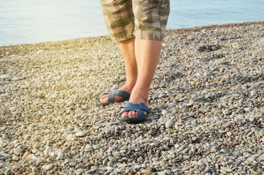 Men's feet in flip-flops and shorts, a man standing on the beach on a pebble beach, sunrise in the morning.