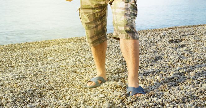 Men's feet in flip-flops and shorts, a man standing on the beach on a pebble beach, sunrise in the morning.