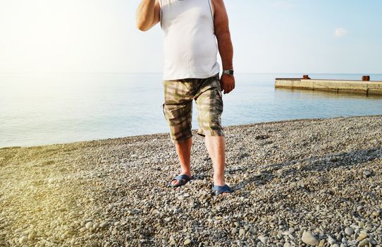 Men's feet in flip-flops and shorts, a man standing on the beach on a pebble beach, sunrise in the morning.