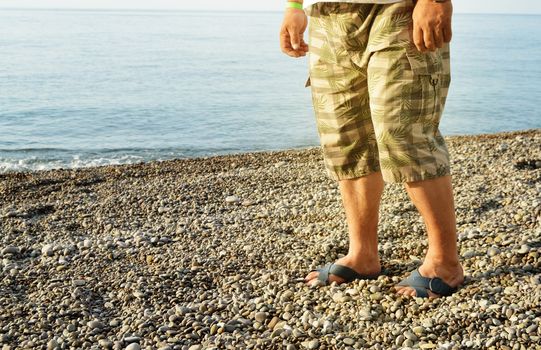 Men's feet in flip-flops and shorts, a man standing on the beach on a pebble beach, sunrise in the morning.