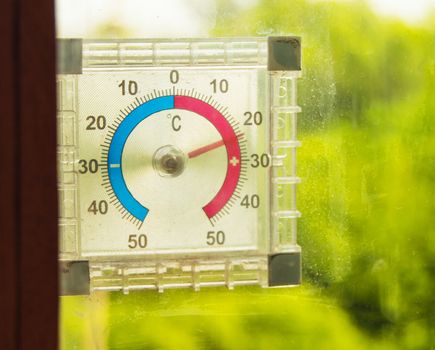 Street thermometer hanging on the window and shows the temperature of the air in the summer against green trees