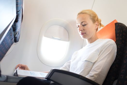 Woman reading in flight magazine on airplane. Female traveler reading seated in passanger cabin. Sun shining trough airplane window.
