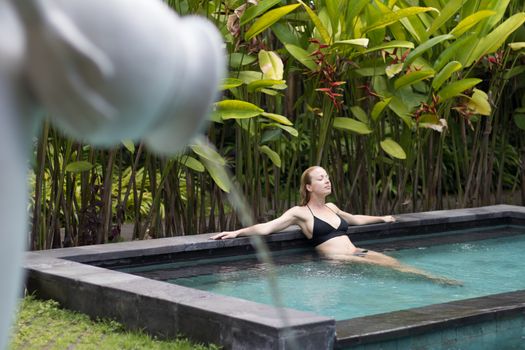 Sensual young woman relaxing in outdoor spa infinity swimming pool surrounded with lush tropical greenery of Ubud, Bali. Wellness, natural beauty and body care concept.