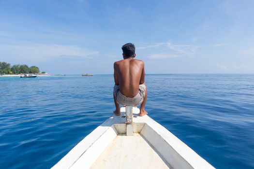 Unrecognizable Local Sporty Guy Sitting Topless at the Bow of Traditional White Wooden Sail Boat, Enjoying the Breeze, Looking At Beautiful Blue Sea of Gili Islands near Bali, Indonesia.