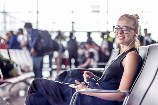 Casual sporty young blond female traveler using her cell phone while waiting to board a plane at the departure gates at the asian airport terminal.