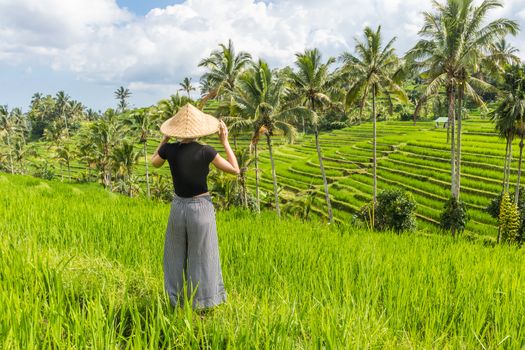 Relaxed fashionable caucasian female tourist wearing small backpack and traditional asian paddy hat looking at beautiful green rice fields and terraces on Bali island.