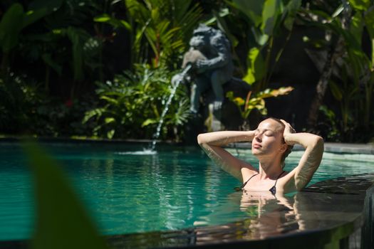 Sensual young woman relaxing in outdoor spa infinity swimming pool surrounded with lush tropical greenery of Ubud, Bali. Wellness, natural beauty and body care concept.