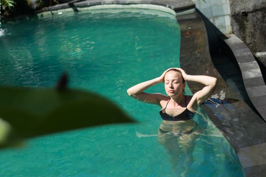 Sensual young woman relaxing in outdoor spa infinity swimming pool surrounded with lush tropical greenery of Ubud, Bali. Wellness, natural beauty and body care concept.