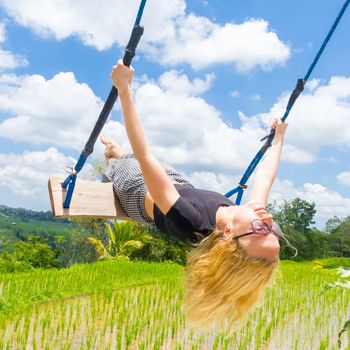Happy caucasian female traveller swinging on wooden swing, enjoying summer vacation in pristine green nature at Jatiluwih rice terraces on Bali, Indonesia. Freedom and life enjoying concept.