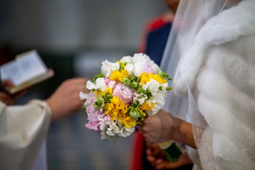 The bride holds a wedding bouquet of beautiful flowers in her hand. The bouquet consists of white, yellow and pink flowers. Priest and brides hands is In the blur area. Bouquet of flowers in the hand of the bride during the marriage ceremony.