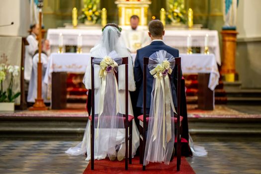 Bride and groom during wedding ceremony in church. Bride and groom preparing for communion on knees at wedding ceremony in church. Chairs decorated with bouquets of flowers and ribbons in church for wedding ceremony.