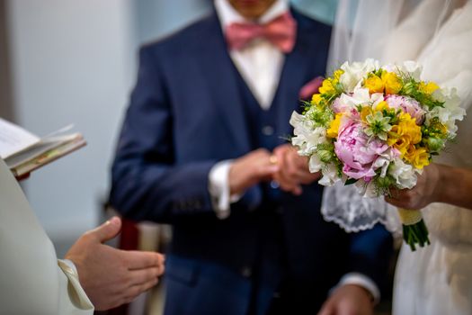The bride holds a wedding bouquet of beautiful flowers in her hand. The bouquet consists of white, yellow and pink flowers. Priest hand and groom is In the blur area. Bouquet of flowers in the hand of the bride during the marriage ceremony.