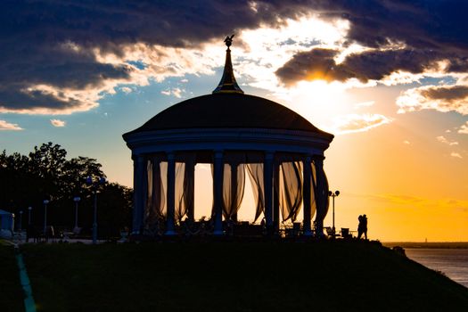 A couple in love hugs and kisses next to a romantic gazebo against the sunset