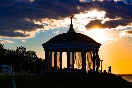 A couple in love hugs and kisses next to a romantic gazebo against the sunset