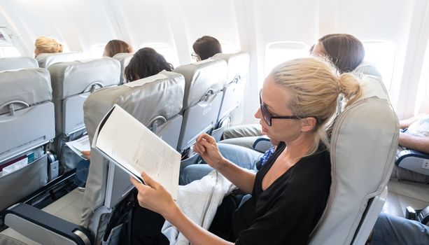 Female traveler reading magazine on airplane during flight. Female traveler reading seated in passanger cabin.