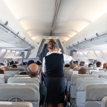 Interior of commercial airplane with flight attandant serving passengers on seats during flight. Stewardess in dark blue uniform walking the aisle.