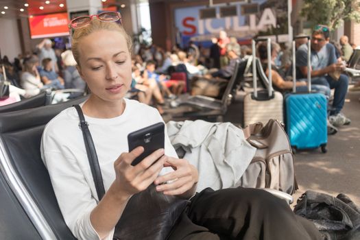 Casual blond young woman reading on her mobile phone while waiting to board a plane at the departure gates at the airport terminal. Travel concept.