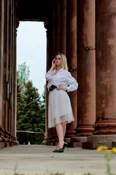 Fashion look's woman. Young woman modern portrait. Young woman dressed in white skirt and shirt posing near the old looking soviet union's architectural building with large pillars and bas-reliefs.