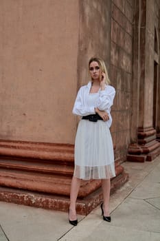 Fashion look's woman. Young woman modern portrait. Young woman dressed in white skirt and shirt posing near the old looking soviet union's architectural building with large pillars and bas-reliefs.