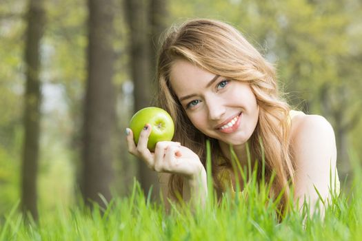 Blonde pretty girl laying on the grass in spring park and smiling holding fresh green apple