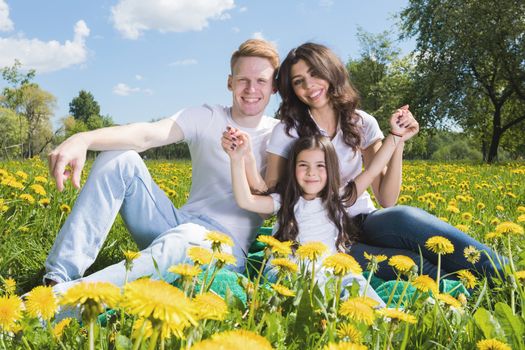 Happy family of parents and daughter sitting on a meadow of dandelion flowers and smiling