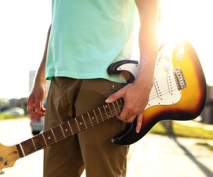 Young hipster in a blue t-shirt with a guitar stands on the road in contrast to sunlight and glare, a summer day outdoors.