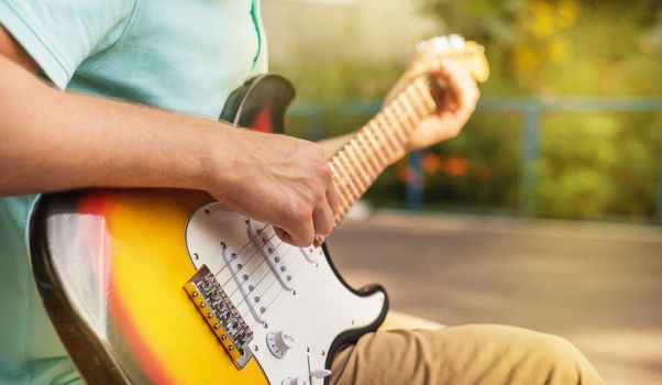 Happy young man hipster sitting playing guitar, close up, summer sunlight, outdoor.