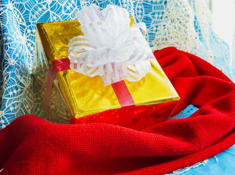 Gift box in gold foil package with white bow and red knitted scarf on beautiful blue Christmas background, Christmas layout concept.