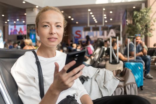 Casual blond young woman reading on her mobile phone while waiting to board a plane at the departure gates at the airport terminal. Travel concept.