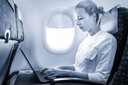 Attractive caucasian female passenger working at modern laptop computer using wireless connection on board of commercial airplane flight. Blue toned greyscale image.