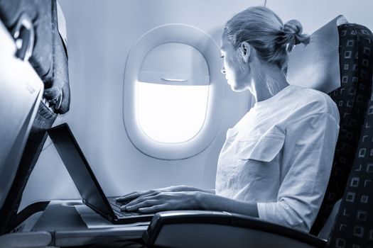 Attractive caucasian female passenger looking through the plain window while working on modern laptop computer using wireless connection on board of commercial flight. Blue toned greyscale image.