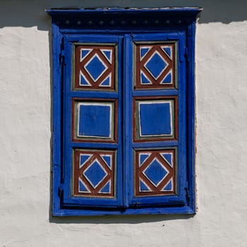 Detail of a traditional village window in Romania painted in strident blue on a white mud wall background.
