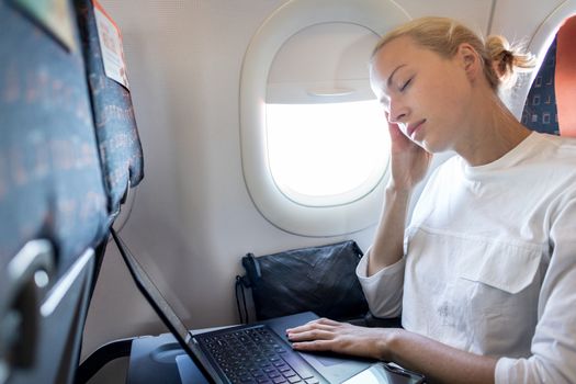 Tired business woman napping on airplane during her business trip woking tasks. Female passenger in flying aircraft sleeps next to the window.