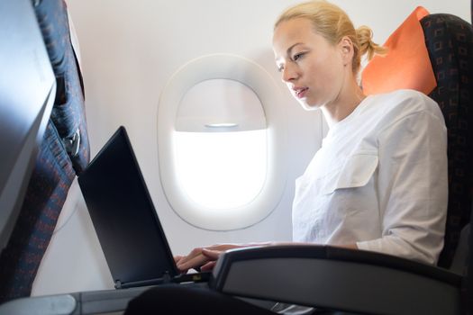 Attractive caucasian female passenger working at modern laptop computer using wireless connection on board of commercial airplane flight.