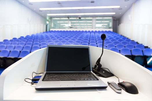 Laptop and microphone on the rostrum in empty conference hall with blue velvet chairs.