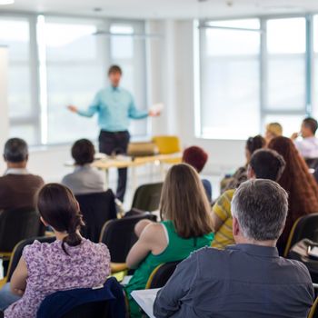 Business and entrepreneurship symposium. Speaker giving a talk at business meeting. Audience in conference hall. Rear view of unrecognized participant in audience.