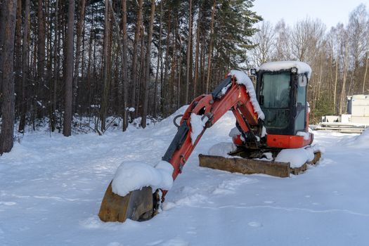 A red excavator near a forest is covered by snow, as the construction site is sitting idle due to the low temperature, snow and ice.