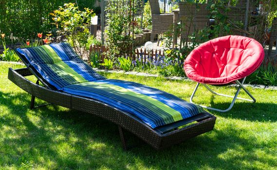 A blue and green colored sun lounger and a red round red folding moon chair in a green garden standing in the sun during spring time..