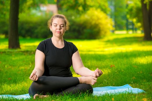 flexible and concentrated woman oversize is meditating on the lawn in the park in the lotus position