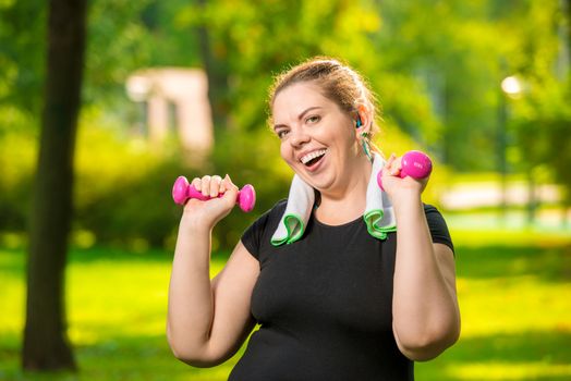 horizontal portrait of a happy plump woman with dumbbells in the park during workout