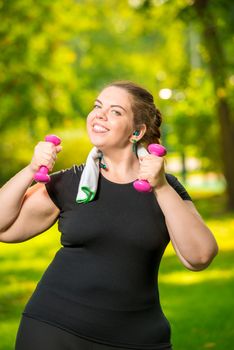 vertical portrait oversize woman in headphones with dumbbells in hand is engaged in sports in the park