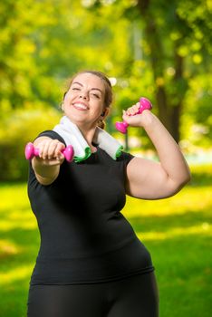 vertical portrait of a happy oversized woman in headphones with dumbbells in her hands playing sports in the park