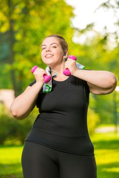 happy plus size model listens to music with headphones and practices dumbbells during a workout in a summer park