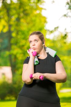 thoughtful plus-size model listens to music with headphones and practices dumbbells during a workout in a summer park