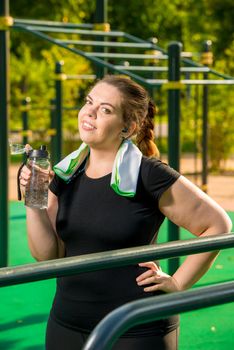 overweight woman with a bottle of clear water rests after training in the park at the simulator