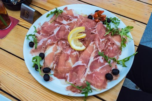 Plate of slice of Parma ham with black olives and lettuce on a wooden table